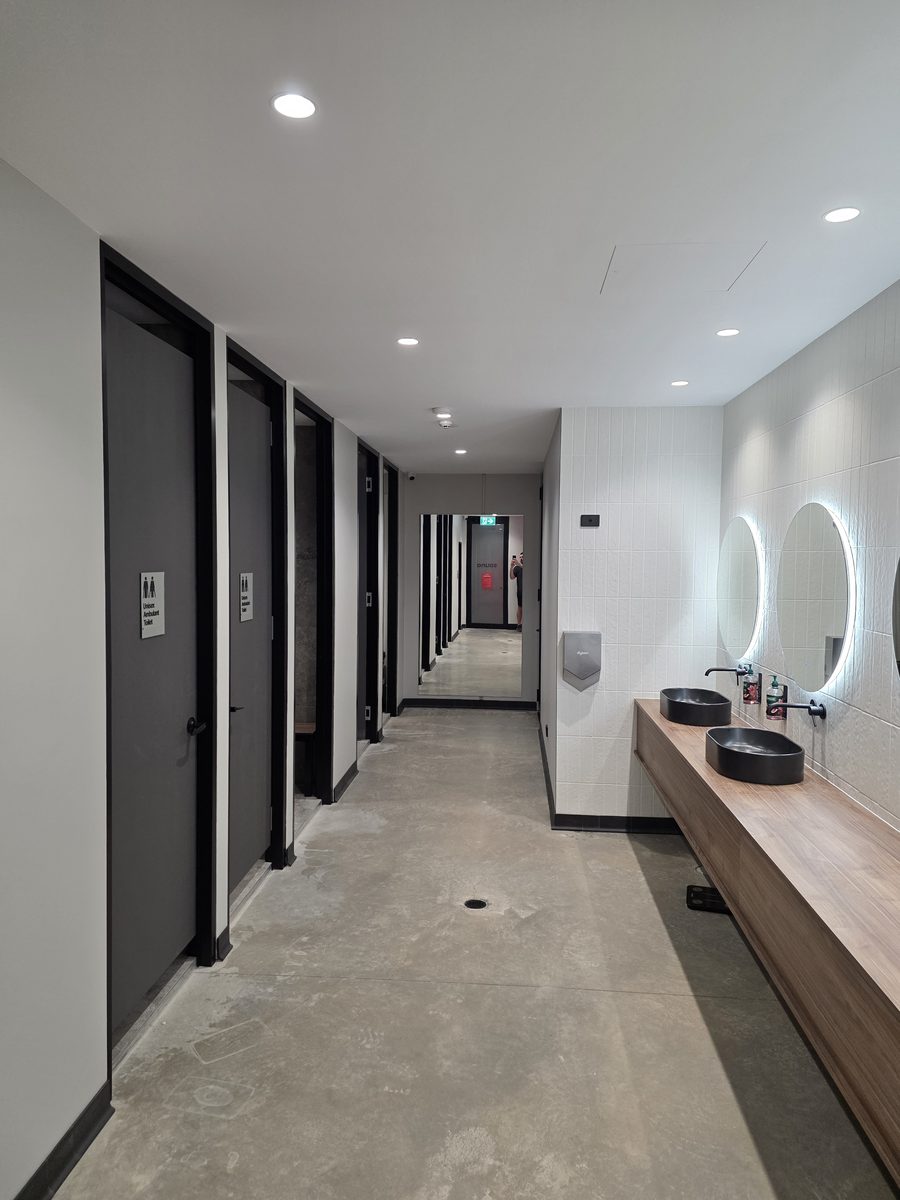 Amenities hallway with cubicles, back-lit oval mirrors and downlights